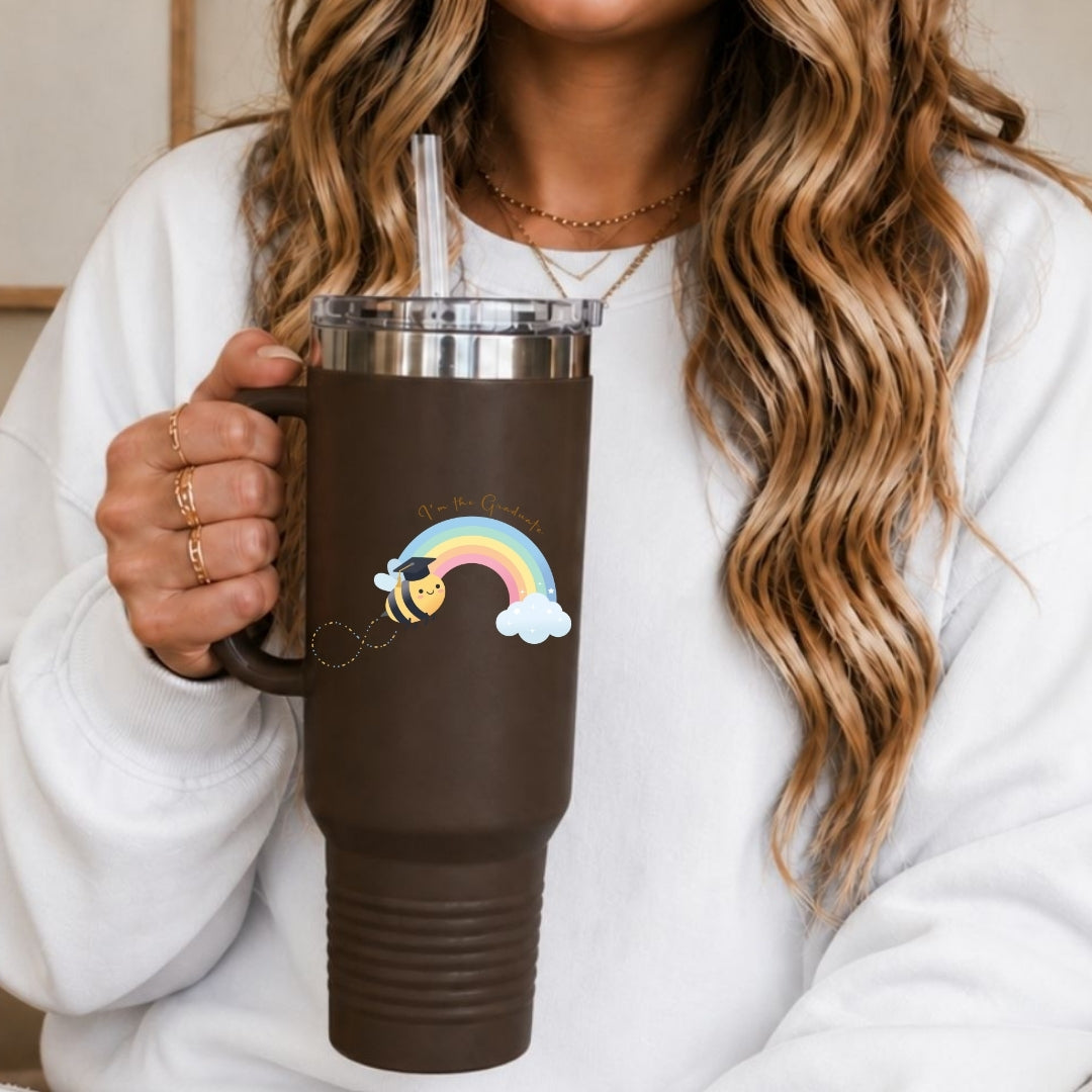 A woman in a white sweatshirt holds a dark brown insulated travel tumbler with a handle, clear straw, and a colorful rainbow bee graphic on the front.
