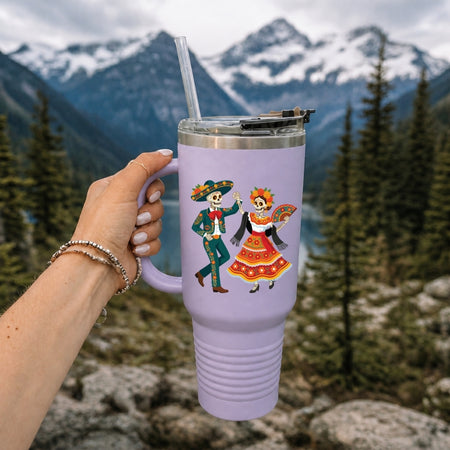 A hand holds a lavender insulated tumbler with a clear straw in front of a mountain and forest background. The tumbler has a colorful Day of the Dead sugar skull illustration on the front.