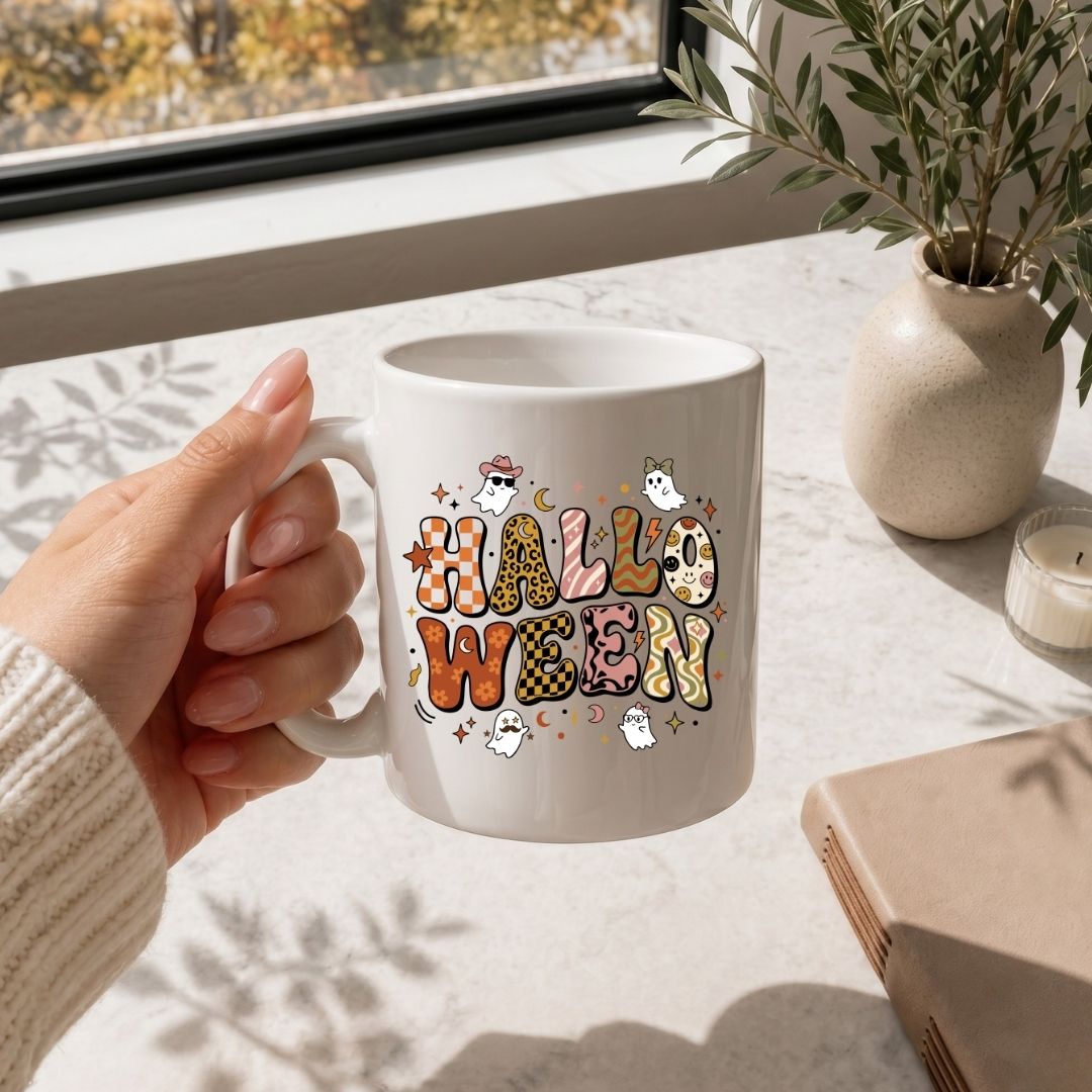 A hand holding a white ceramic mug with a colorful 'HALLOWEEN' graphic featuring pumpkins, ghosts, and stars, set on a light marble tabletop near a window with a plant and candle.