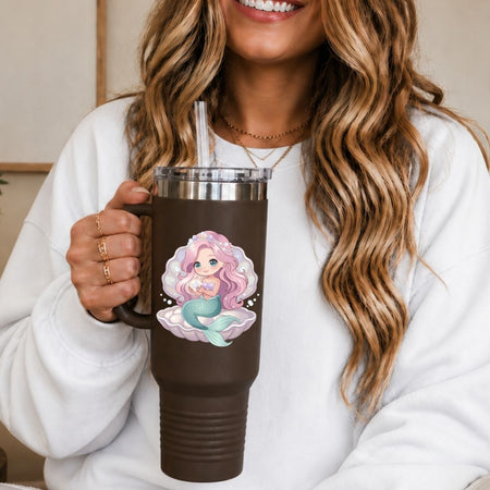 A woman in a white sweatshirt holds a dark brown insulated tumbler with a clear lid and straw. The tumbler features a printed illustration of a mermaid with pink hair and a teal tail on a white cloud.