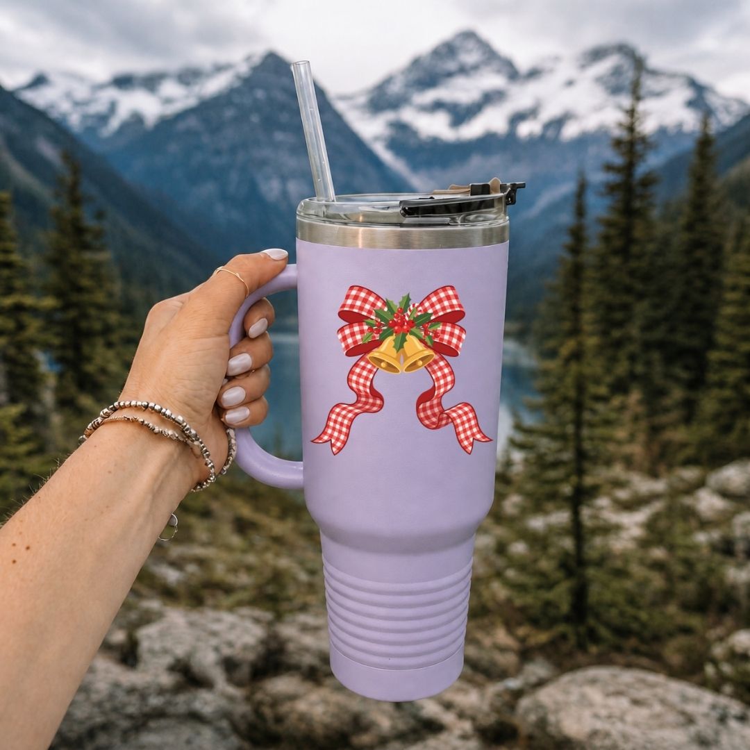 A lavender insulated tumbler with a red and white gingham bow and gold bells graphic, held by a hand against a blurred mountain and forest background.