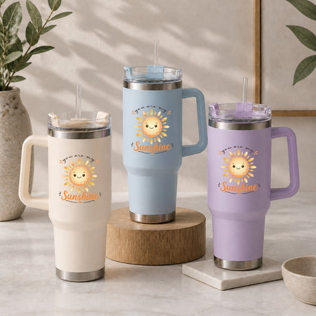 Three stainless steel tumblers in cream, light blue, and light purple, each decorated with the same happy cartoon sun graphic and the text "you are my Sunshine".