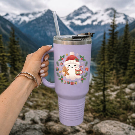 A lavender insulated tumbler with a handle, clear straw, and lid, held by a hand against a blurred mountain and forest background. The tumbler features a cute cartoon snowman wearing a red Santa hat surrounded by a holiday wreath.
