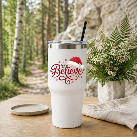 White travel tumbler with a black straw, featuring a holiday "Believe" design, placed on a wooden table near a plant.