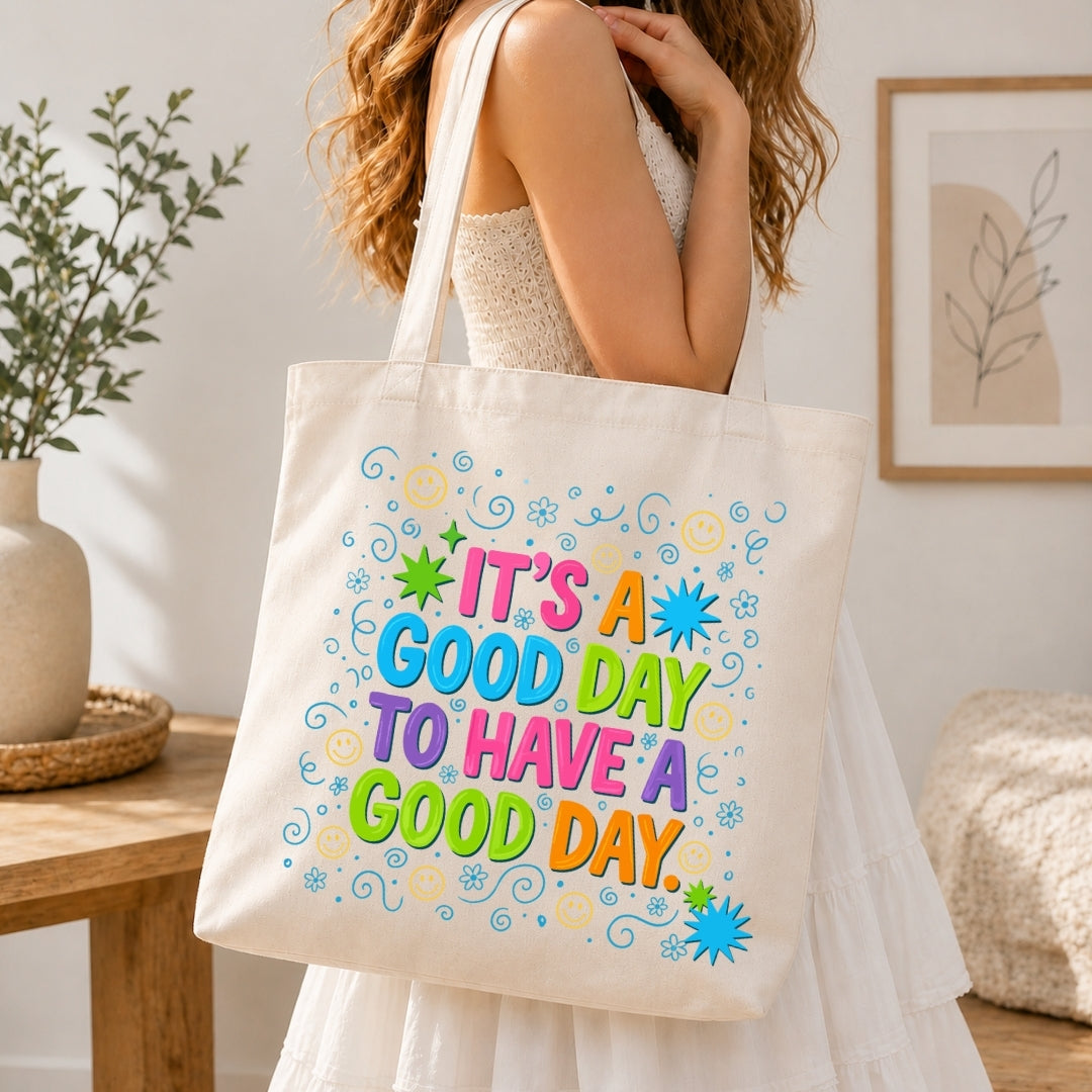 A woman in a white dress carries a cream canvas tote bag with colorful text that reads 'IT'S A GOOD DAY TO HAVE A GOOD DAY.' in a bright, modern room setting.