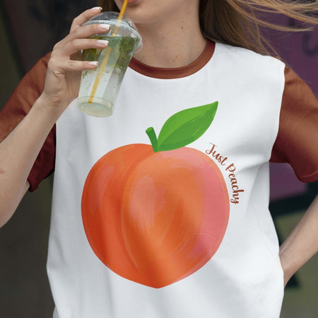 Person wearing a white t-shirt with a peach graphic and 'Just Peachy' text, holding a drink.