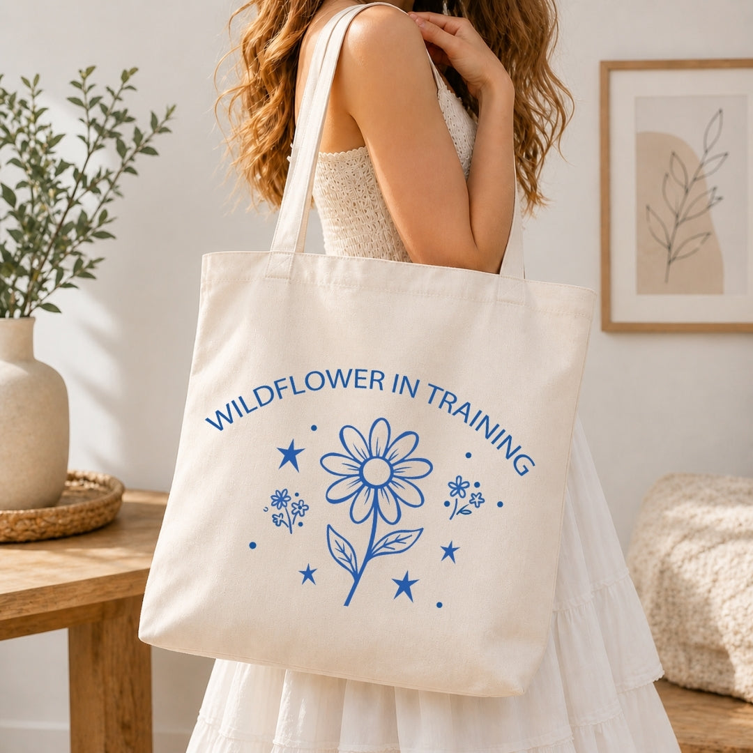 Woman in a white tiered dress carrying a cream-colored canvas tote bag printed with a large blue "Wildflower in Training" retro daisy graphic over her shoulder.