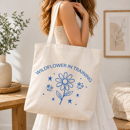 Woman in a white tiered dress carrying a cream-colored canvas tote bag printed with a large blue "Wildflower in Training" retro daisy graphic over her shoulder.