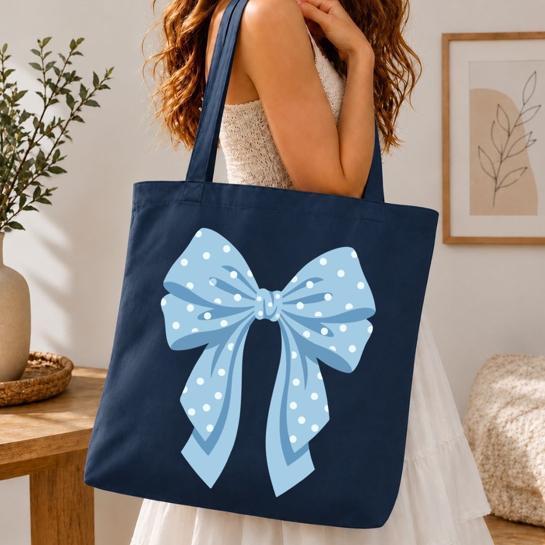 A navy blue canvas tote bag with a large light blue polka-dot bow graphic on the front, carried by a woman in a white dress in a bright indoor room.