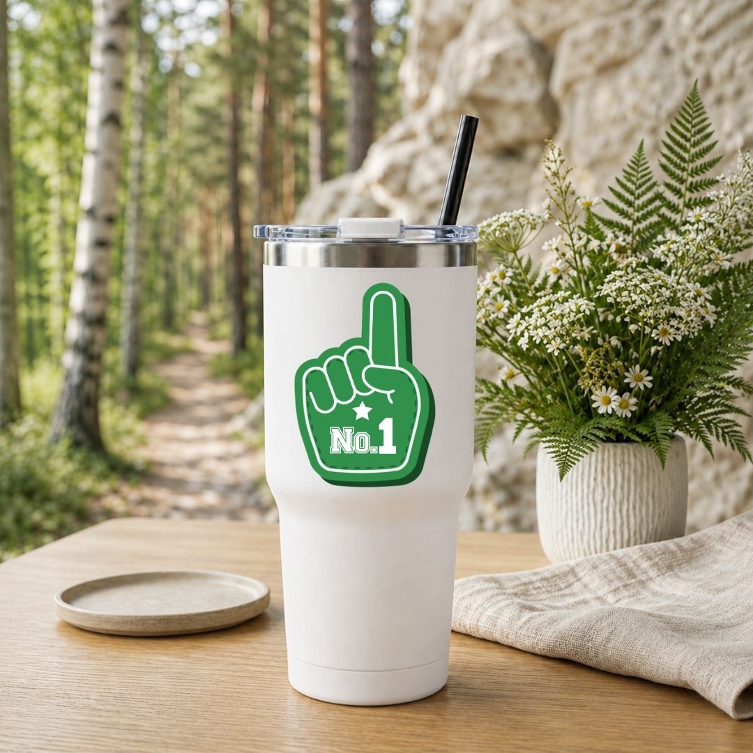 A white insulated tumbler with a clear lid and black straw sits on a wooden table outdoors. The tumbler has a large green foam-finger graphic with the text 'No.