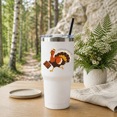 A white insulated tumbler with a clear lid and black straw stands on a wooden table in a forest setting. The tumbler is printed with a cartoon turkey carrying a suitcase and the phrase 'And Poultry Meets the World.