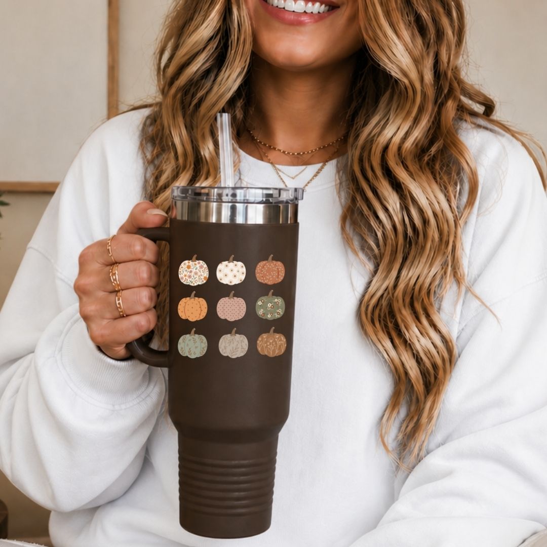 A woman in a white sweatshirt holds a dark brown insulated tumbler with a handle, clear lid, and straw. The tumbler is decorated with a grid of small fall pumpkins in muted orange, beige, and sage green tones.