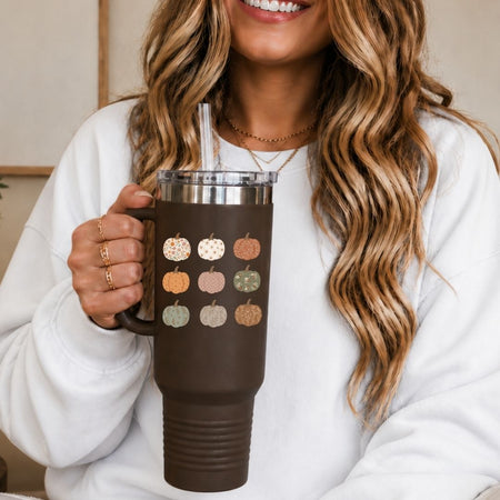 A woman in a white sweatshirt holds a dark brown insulated tumbler with a handle, clear lid, and straw. The tumbler is decorated with a grid of small fall pumpkins in muted orange, beige, and sage green tones.