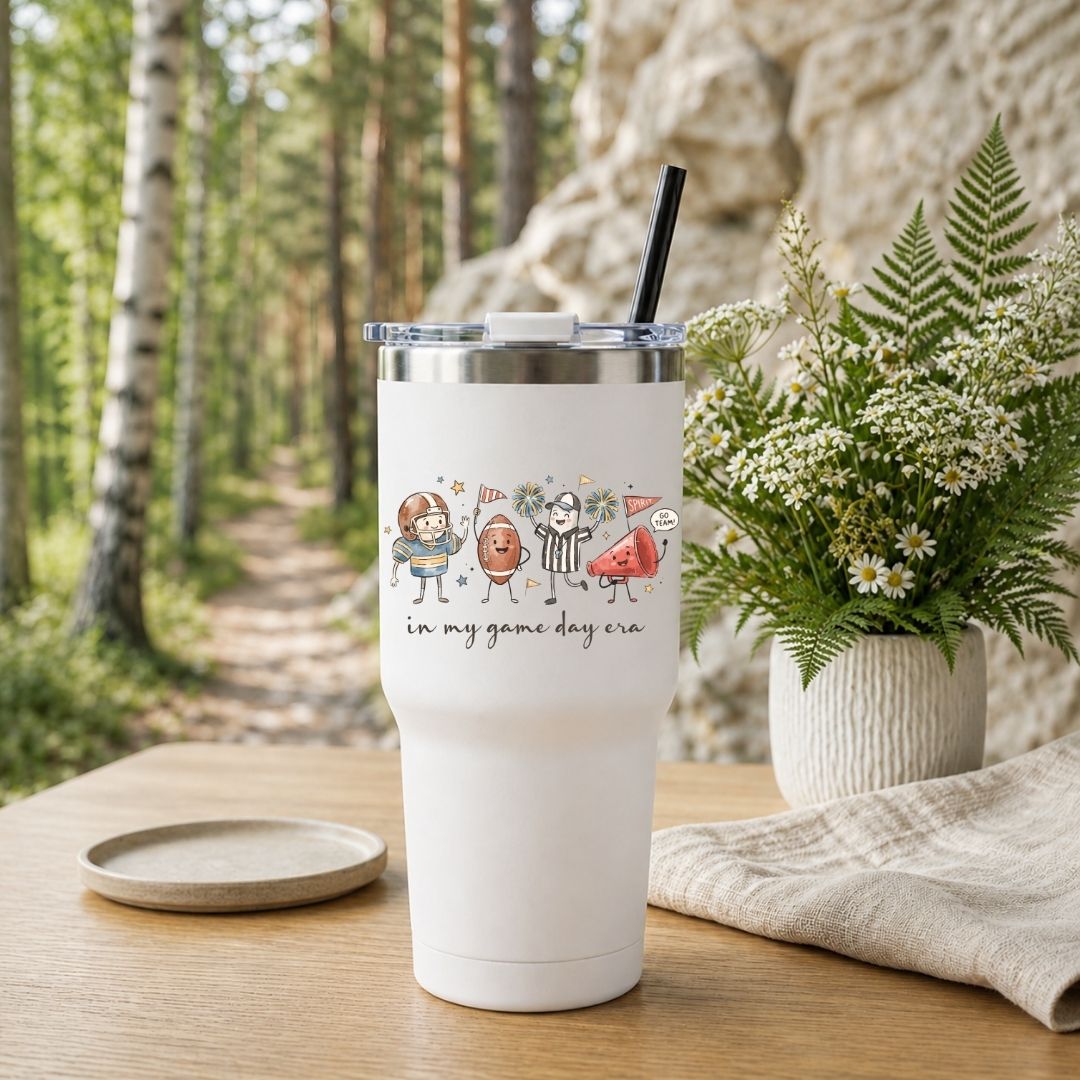 A white insulated tumbler with a clear lid and black straw sits on a wooden table outdoors. The tumbler has a printed cartoon graphic of sports figures and the text 'in my game day era.