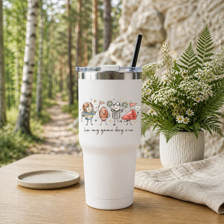 A white insulated tumbler with a clear lid and black straw sits on a wooden table outdoors. The tumbler has a printed cartoon graphic of sports figures and the text 'in my game day era.