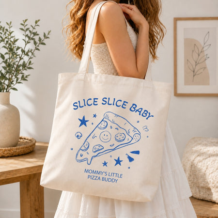 Woman in a white tiered dress carrying a cream-colored canvas tote bag printed with a large blue retro pizza slice illustration over her shoulder.