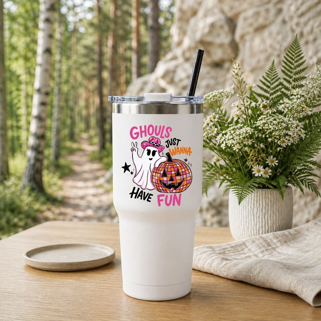 A white stainless steel travel tumbler with a black straw, featuring the Ghouls Just Wanna Have Fun design, placed on a wooden table outdoors with a forest background.