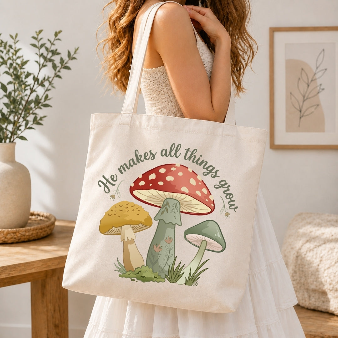 A woman in a white dress carries a cream canvas tote bag featuring a colorful illustration of three mushrooms and the text 'He makes all things grow'. The scene is set in a warm, softly lit room with a wooden table, potted plant, and framed wall art.