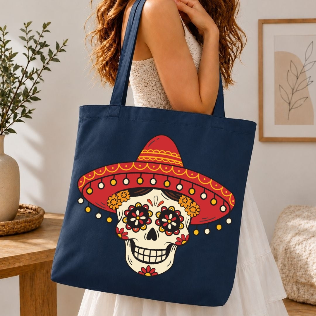 A woman wearing a white dress carries a navy blue canvas tote bag featuring a colorful sugar skull with a red sombrero design, set in a cozy indoor environment.