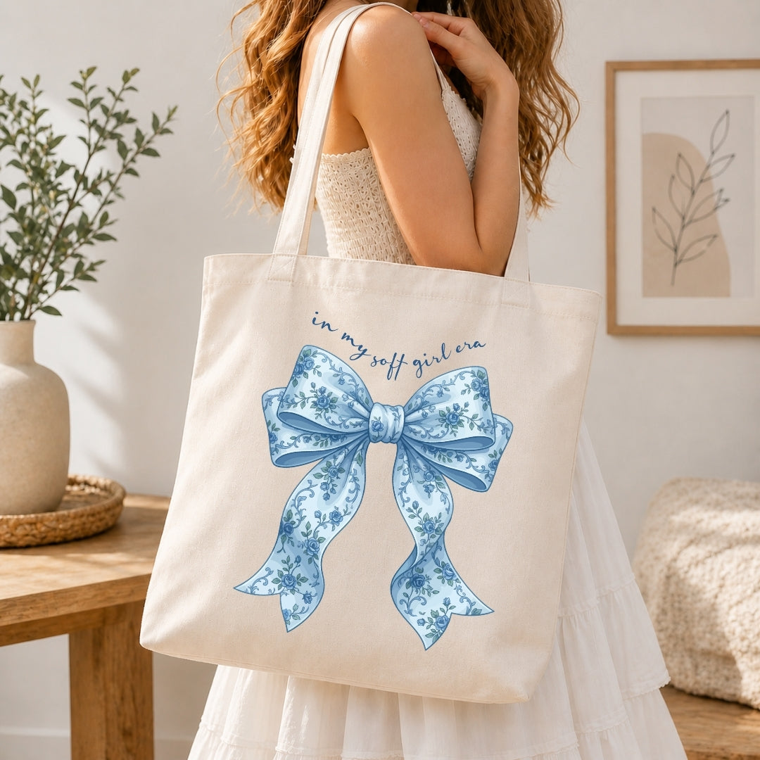 A woman holding a cream-colored tote bag printed with a blue floral bow graphic and the text "in my soft girl era."