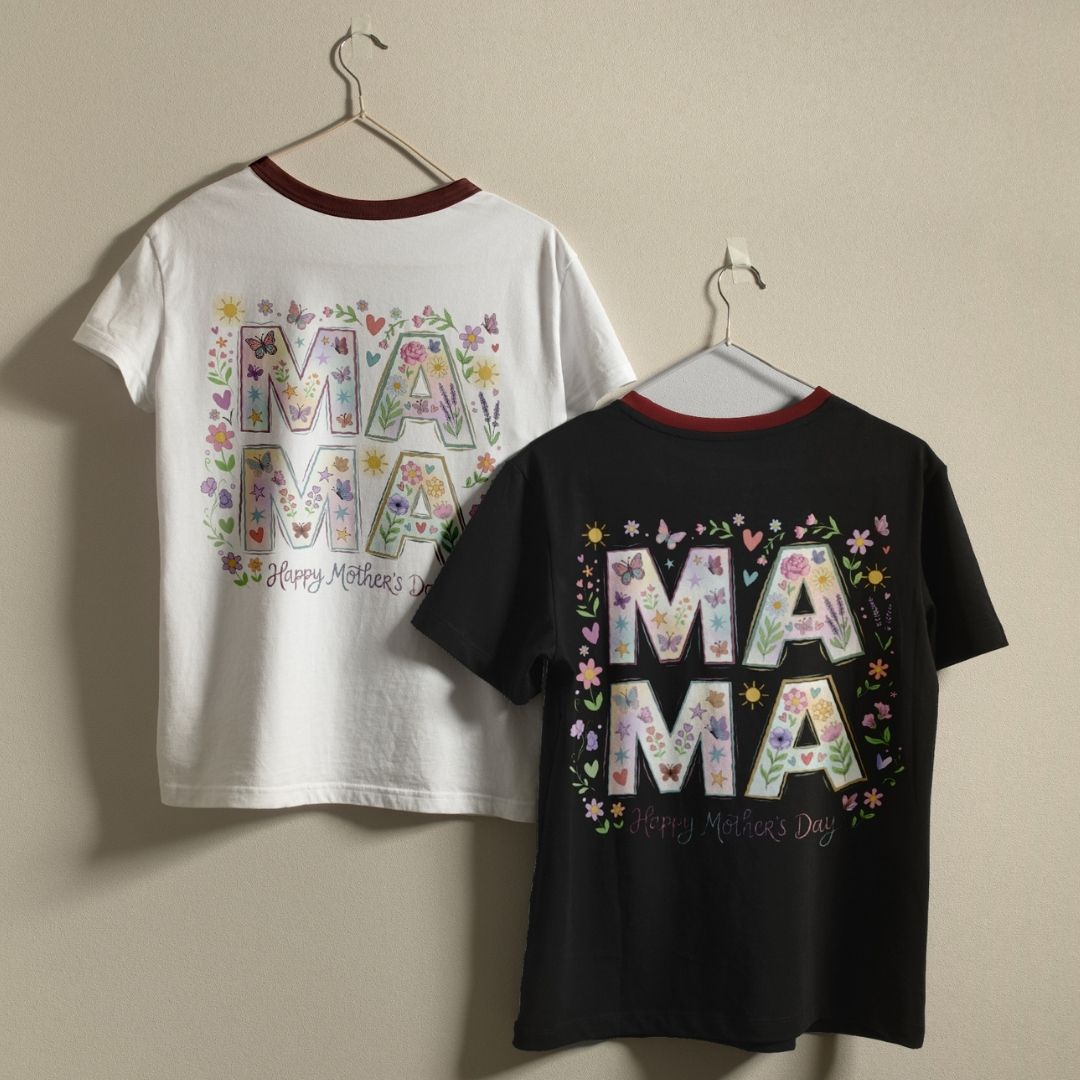 Two t-shirts, one white and one black, with floral 'MAMA' text design on hangers against a beige wall.