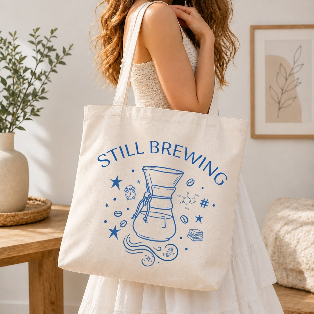 Woman in a white tiered dress carrying a cream-colored canvas tote bag printed with a large blue retro "Still Brewing" coffee maker graphic over her shoulder.