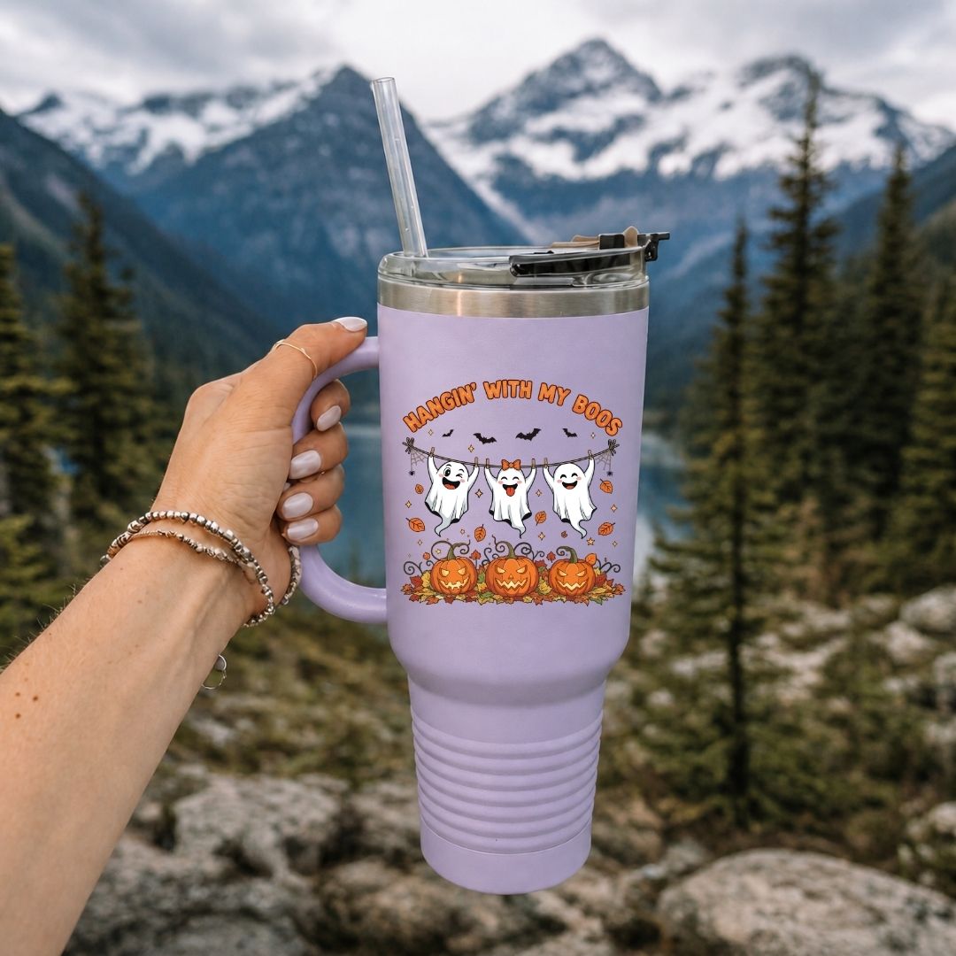 A hand holding a light purple tumbler with a straw, featured in an outdoor mountain setting, printed with the "HANGIN' WITH MY BOOS" cute ghost illustration.