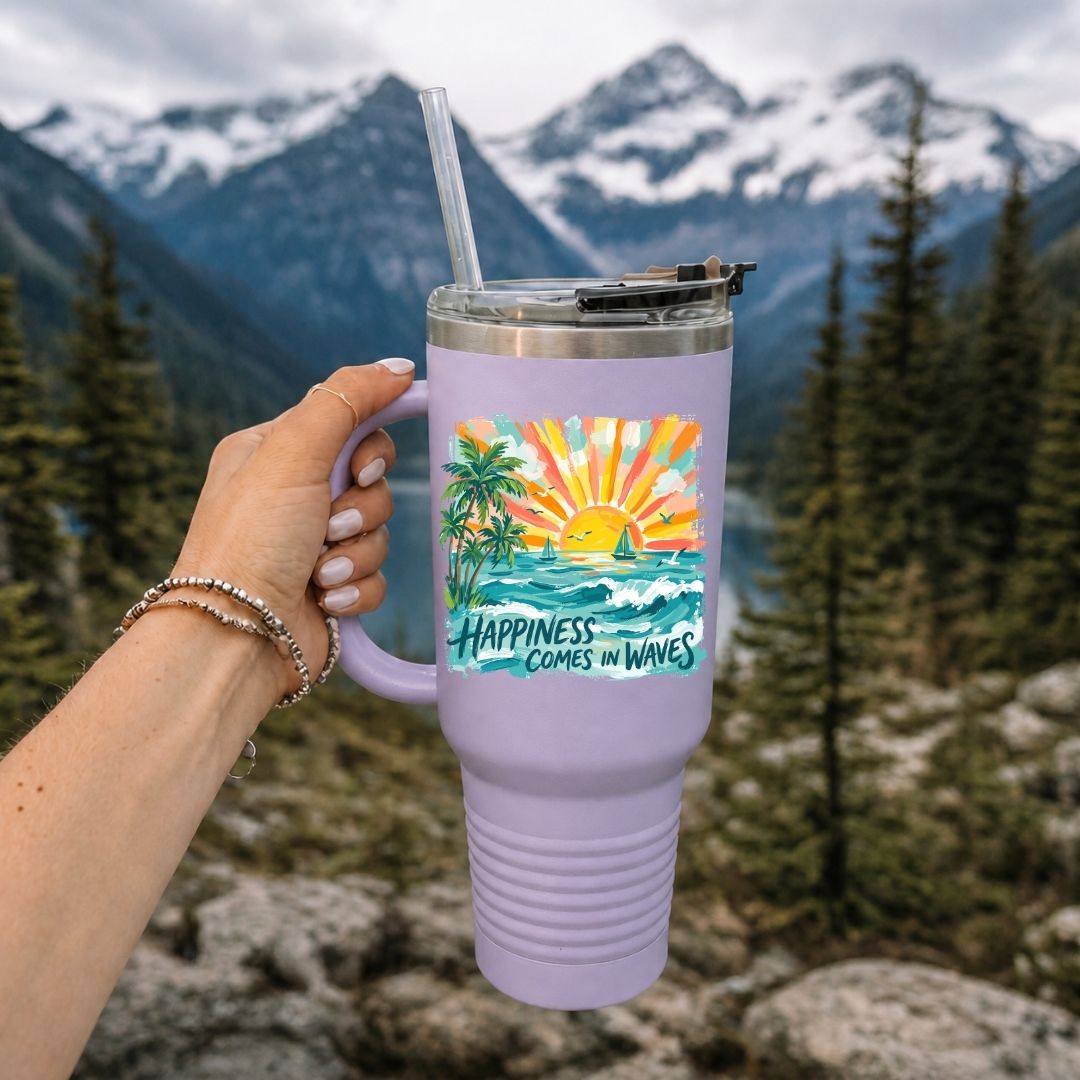 A lavender insulated tumbler with a handle, clear straw, and lid, held by a hand against a mountain and forest background. The tumbler features a colorful tropical sunset graphic with the text 'HAPPINESS COMES IN WAVES'.