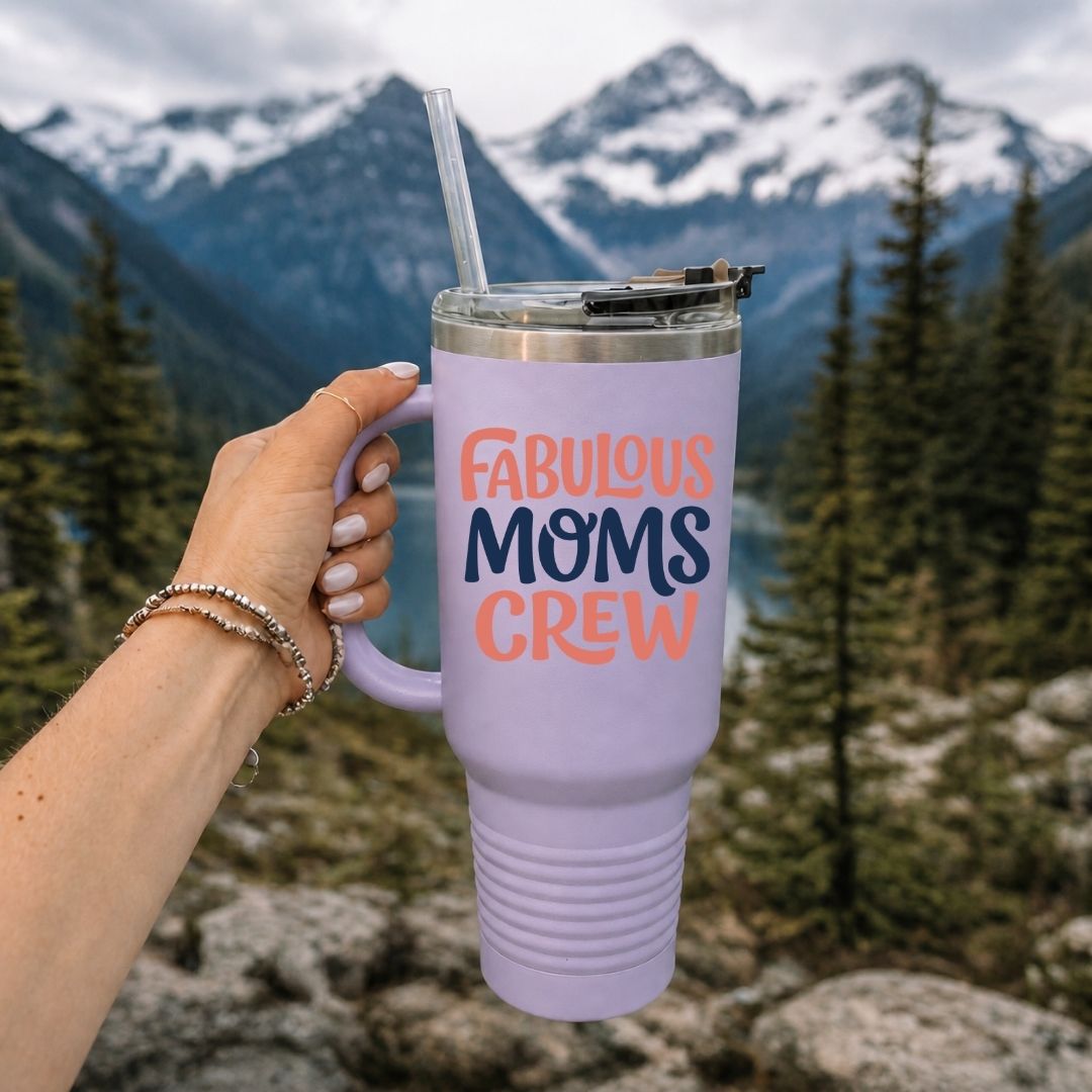 A hand holds a lavender insulated tumbler with a clear straw in front of a mountain and forest background. The tumbler has the words “FABULOUS MOMS CREW” printed on it in coral and navy lettering.