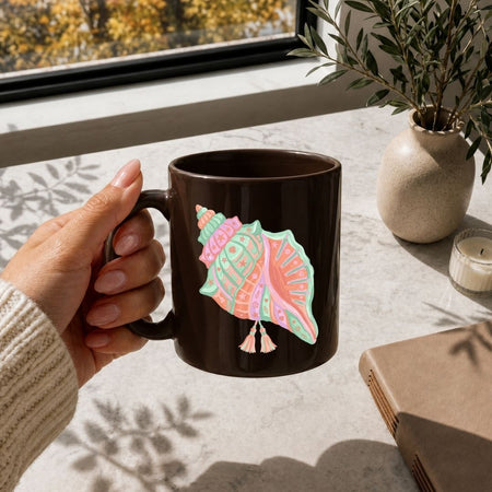 Hand holding a brown mug with a colorful seashell design on a table with a notebook and plant.