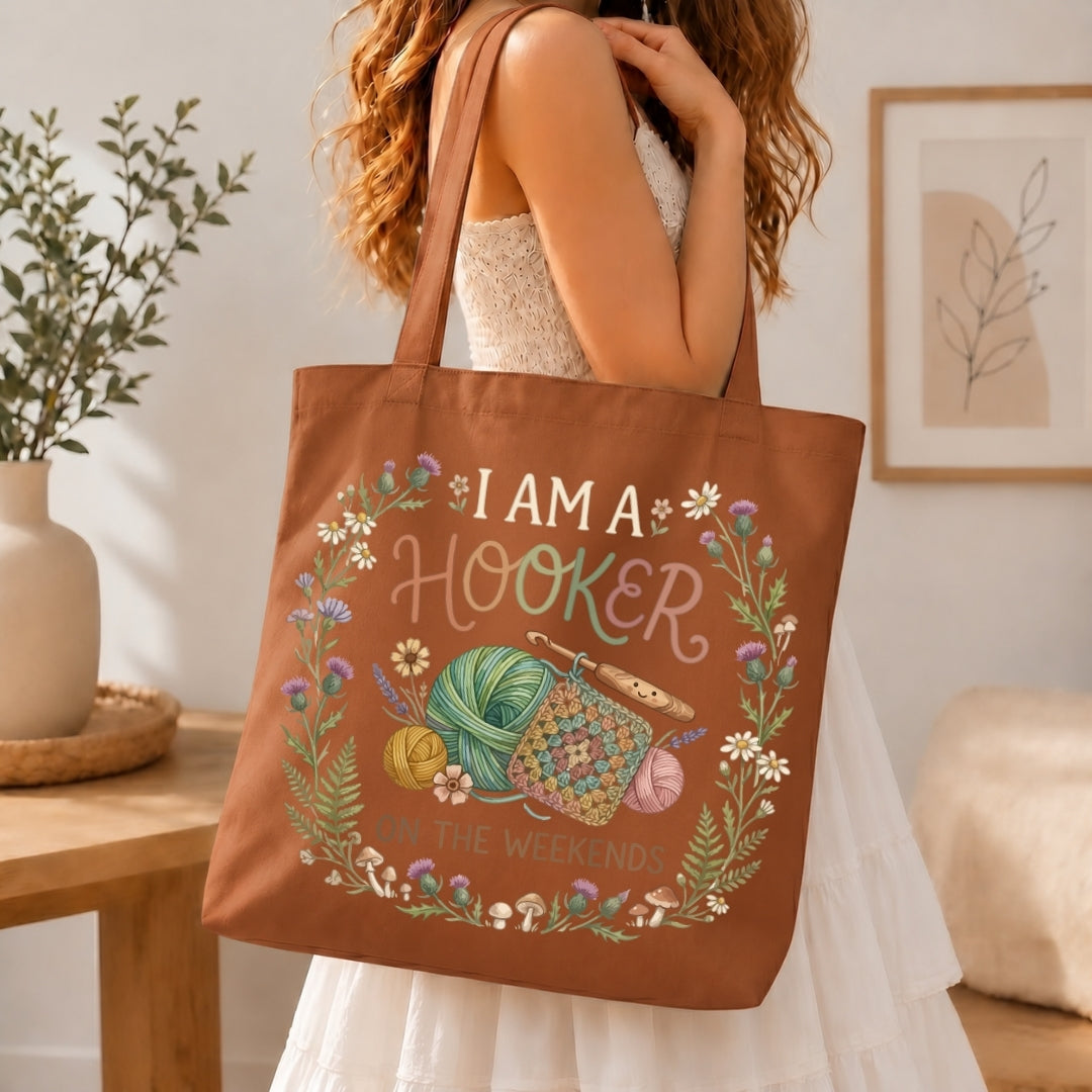 A woman in a white dress holds a brown canvas tote bag with a colorful knitting-themed graphic that reads 'I AM A HOOKER' and 'KNOCKING THE WEEKENDS'. The scene is set in a cozy, softly lit room with a wooden table, potted plant, and framed wall art.