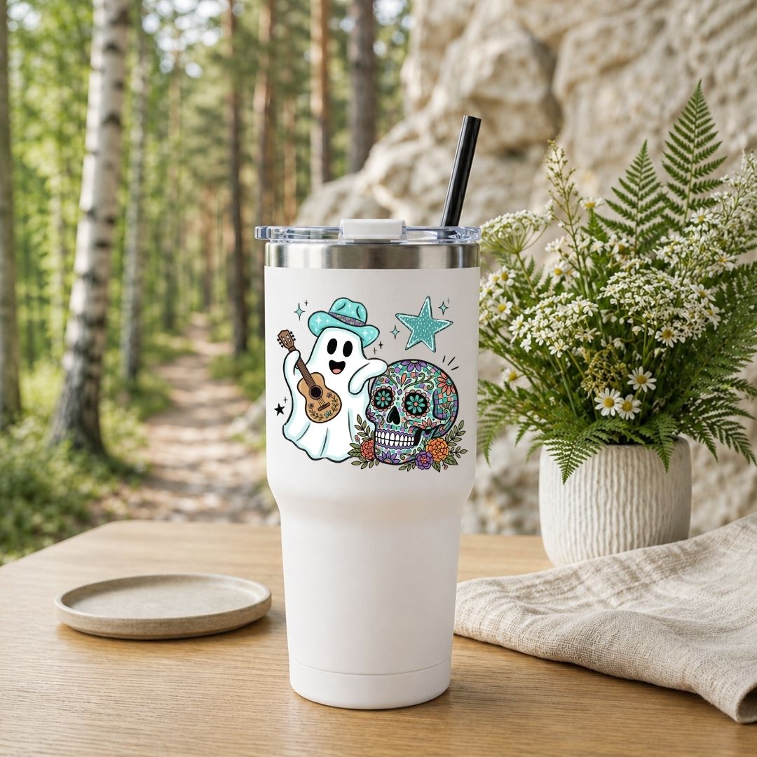 A white insulated tumbler with a clear lid and black straw stands on a wooden table outdoors. The tumbler is printed with a colorful Day of the Dead style design featuring a ghost playing a ukulele next to a decorated sugar skull and star.