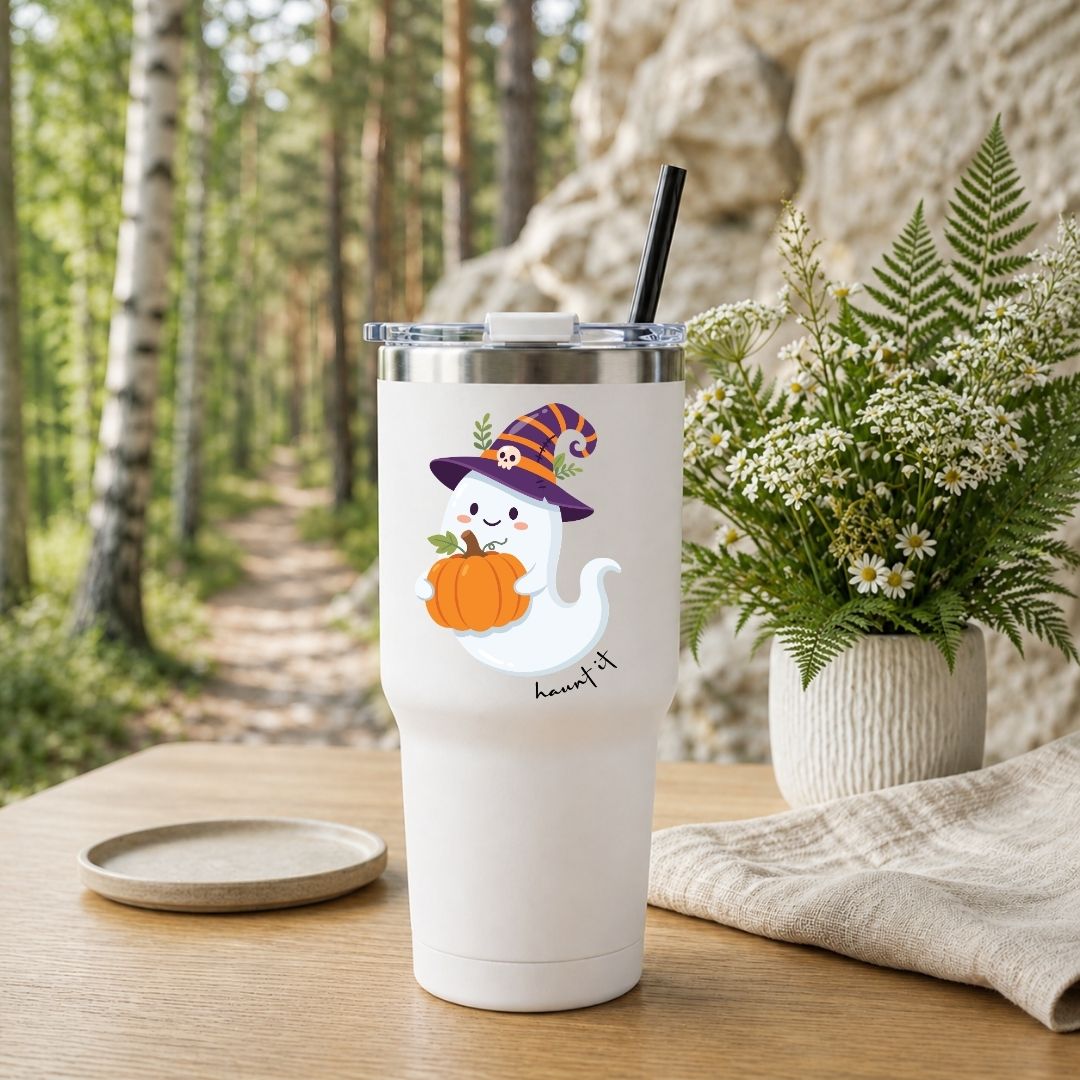 A white insulated tumbler with a lid and black straw sits on a wooden table in a forest setting, decorated with a cute cartoon ghost wearing a witch hat and holding a pumpkin. A small matching coaster is placed beside it, with a potted fern and stone wall in the background.