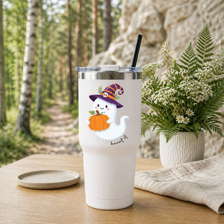 A white insulated tumbler with a lid and black straw sits on a wooden table in a forest setting, decorated with a cute cartoon ghost wearing a witch hat and holding a pumpkin. A small matching coaster is placed beside it, with a potted fern and stone wall in the background.