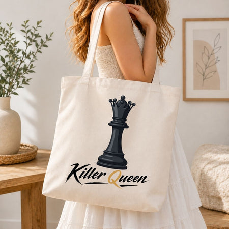 A woman in a white dress carries a cream canvas tote bag featuring a black chess queen graphic and the words 'Killer Queen' in black and gold script, set against a warm, softly lit indoor background.