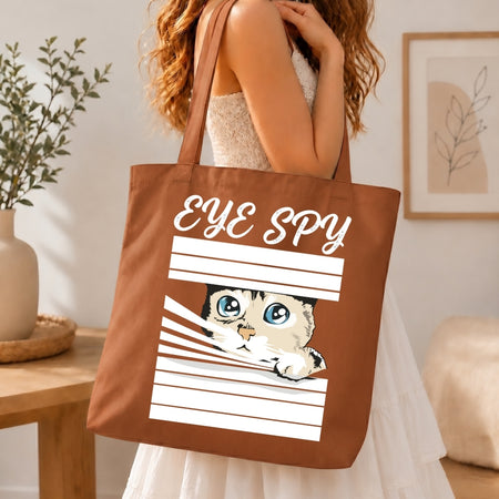 Woman in a white tiered dress carrying a brown canvas tote bag printed with a large white "EYE SPY" window blinds peeking cat graphic over her shoulder.