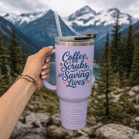 A lavender insulated travel tumbler with a handle, clear straw, and metal lid, held by a hand against a mountain and forest background. The tumbler has the phrase 'Coffee, Scrubs, and Saving Lives' printed on it in dark blue script with small pink and orange accents.