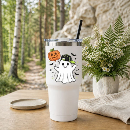 A white insulated tumbler with a Halloween graphic of a ghost and pumpkin, shown on a wooden table with a blurred forest and rock background, next to a potted fern and a small dish.