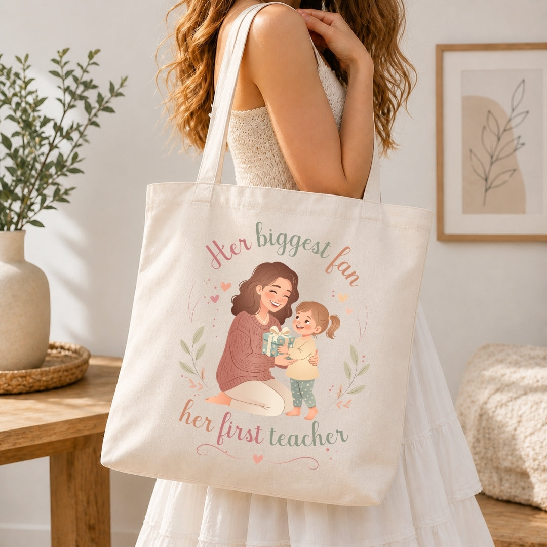 Woman carrying a cream-colored canvas tote bag over her shoulder featuring the mother-daughter graphic illustration and Her biggest fan, her first teacher text.