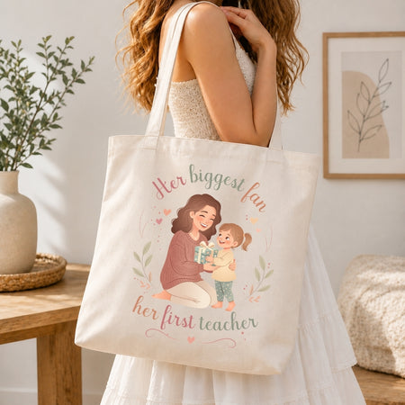 Woman carrying a cream-colored canvas tote bag over her shoulder featuring the mother-daughter graphic illustration and Her biggest fan, her first teacher text.