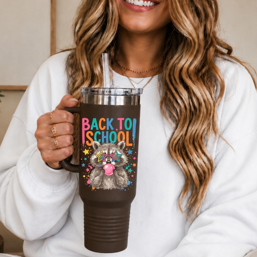 A woman in a white sweatshirt holds a dark brown insulated tumbler with a clear lid and straw. The tumbler features a colorful graphic of a raccoon with sunglasses and the text 'BACK TO SCHOOL' in rainbow letters.