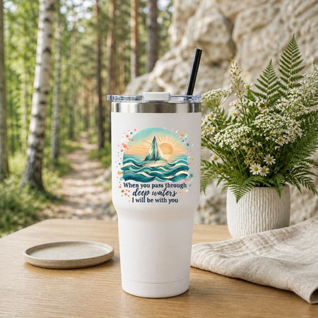 A white insulated tumbler with a clear lid and black straw sits on a wooden table in a softly blurred outdoor setting with trees, a stone wall, a small plate, and a potted plant. The tumbler has a printed ocean-wave sunset design with the text, 'When you pass through deep waters, I will be with you.
