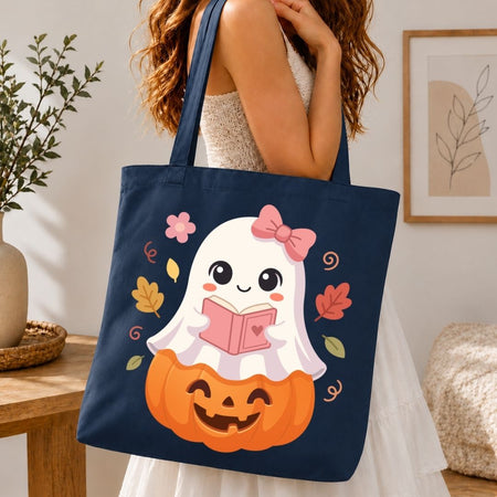 A navy blue canvas tote bag with a cute cartoon ghost and pumpkin graphic, carried by a woman in a white dress in a cozy indoor setting.