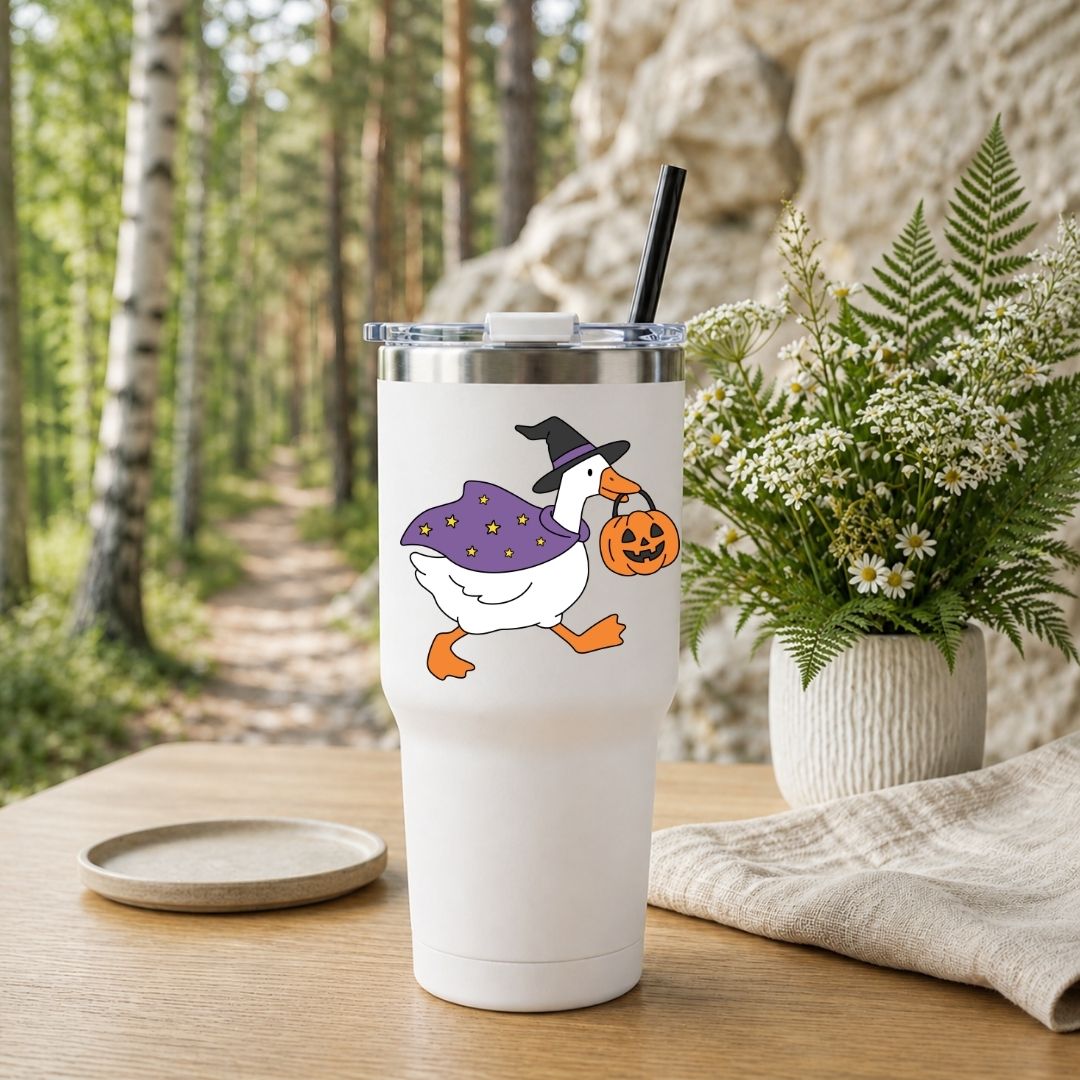 A white insulated tumbler with a clear lid and black straw stands on a wooden table in a forest setting. The tumbler is printed with a cartoon white goose wearing a black witch hat and purple cape while carrying a jack-o'-lantern.