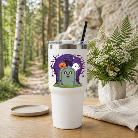 A white insulated tumbler with a clear lid and black straw sits on a wooden table in a forest setting. The tumbler has a Halloween-themed graphic showing a green tombstone monster, a white ghost, a jack-o'-lantern, and a purple spooky tree.