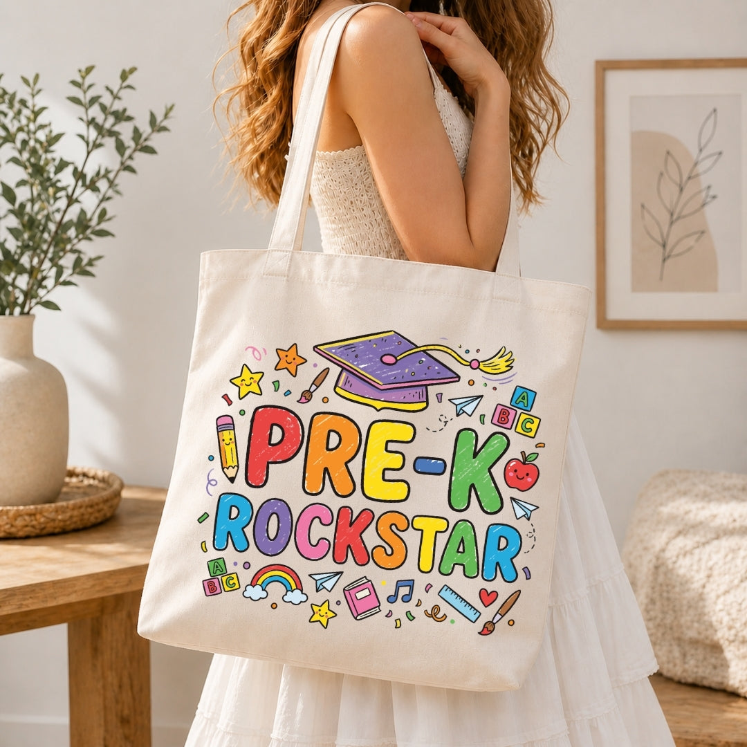 A woman wearing a white sleeveless dress carries a cream canvas tote bag with colorful 'PRE-K ROCKSTAR' lettering and school-themed doodles, set in a bright, modern room.