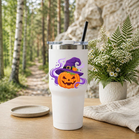 A white insulated tumbler with a lid and black straw stands on a wooden table outdoors. The tumbler has a Halloween graphic of a carved orange pumpkin under a purple witch hat with purple smoke.