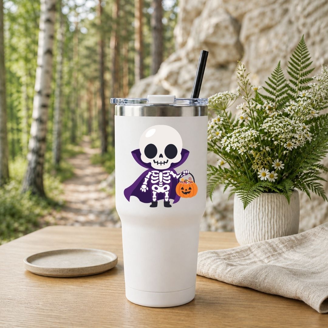 A white insulated tumbler with a clear lid and black straw, featuring a cartoon skeleton wearing a purple cape and holding a pumpkin bucket. The tumbler is set on a wooden table outdoors with a blurred forest and rock background, alongside a small plate and a potted fern.