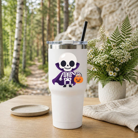 A white insulated tumbler with a clear lid and black straw, featuring a cartoon skeleton wearing a purple cape and holding a pumpkin bucket. The tumbler is set on a wooden table outdoors with a blurred forest and rock background, alongside a small plate and a potted fern.
