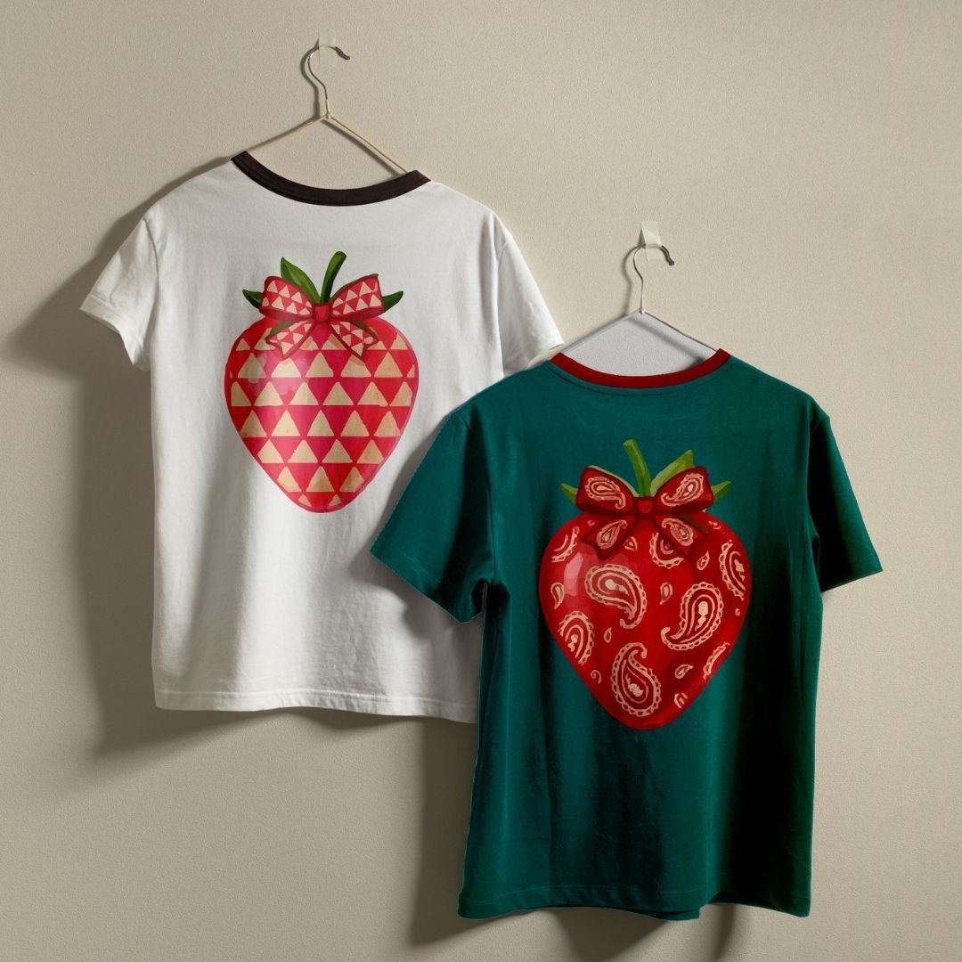 Two children's short-sleeve ringer T-shirts hanging on hangers against a plain beige wall. The left shirt is white with a dark brown collar and features a large red strawberry graphic on the back with pink triangles and a pink bow.
